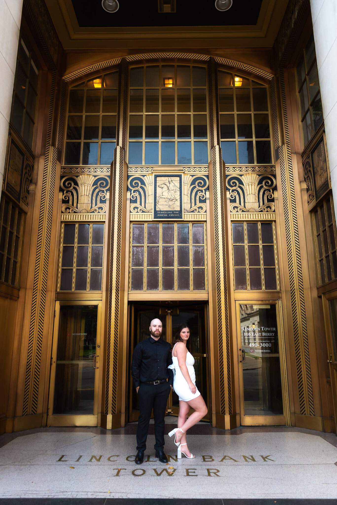Young woman with long brown hair in white dress, standing next to young man with beard in black suit, in front of golden Art Deco doors