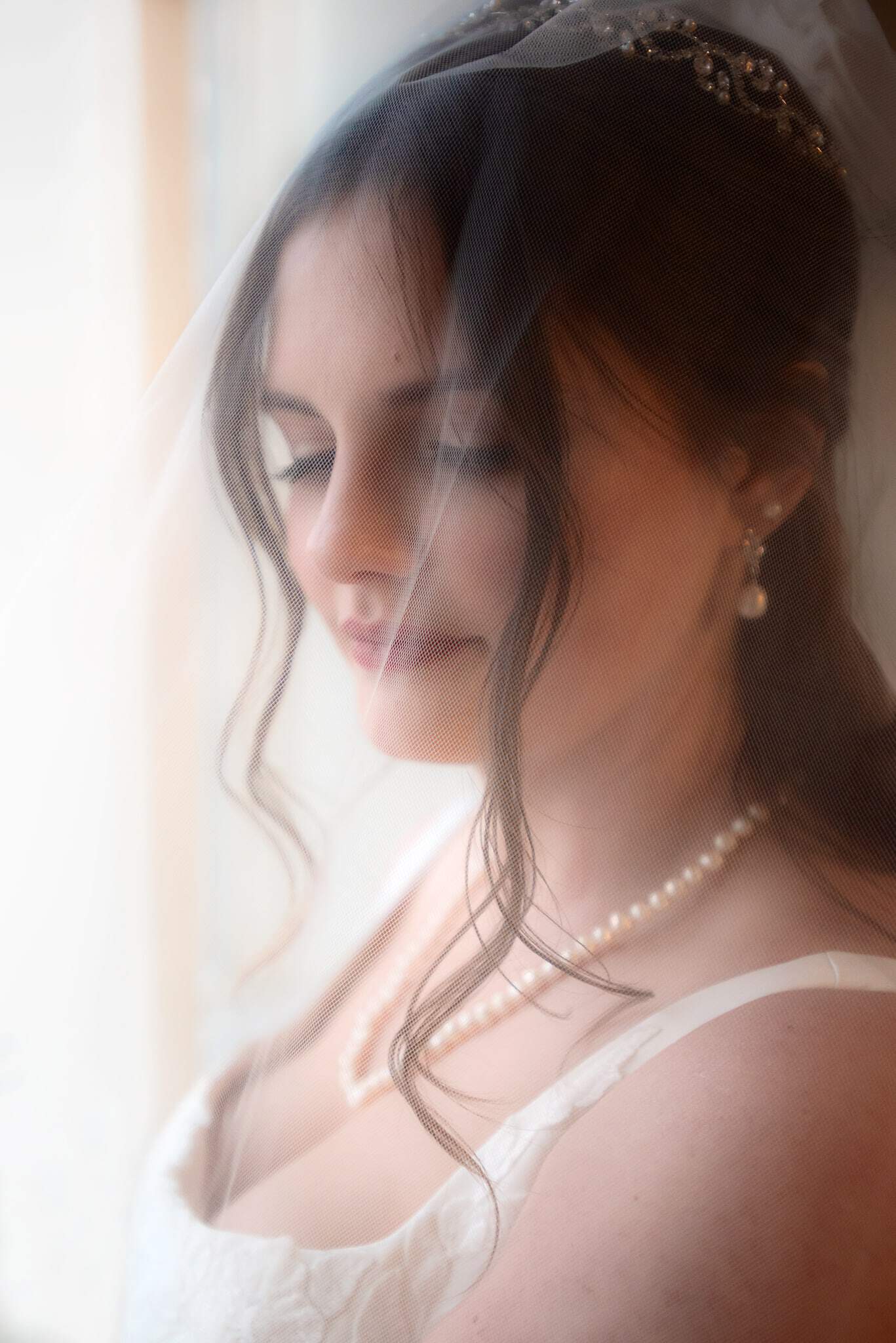 Young woman with long, dark brown hair and tiara, poses for demure portrait with long white veil over face in front of window